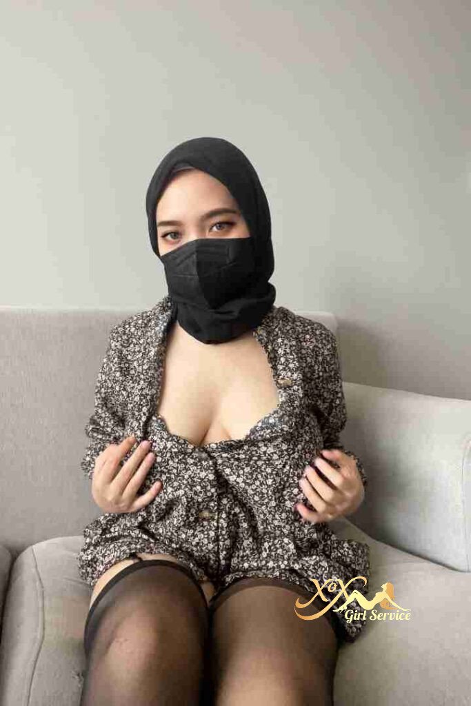 Khayla, Malay woman in hijab and mask, floral top, stockings, sitting on sofa.