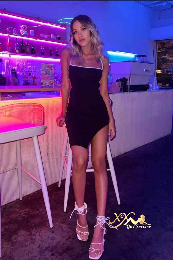 Bella, Thai escort in black dress and white heels, standing in bar with neon lights.